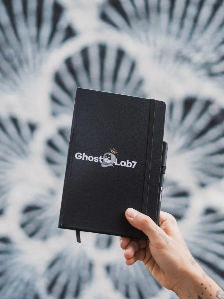 Mão segurando o caderno preto da Ghost Lab7 diante de um mural em preto e branco